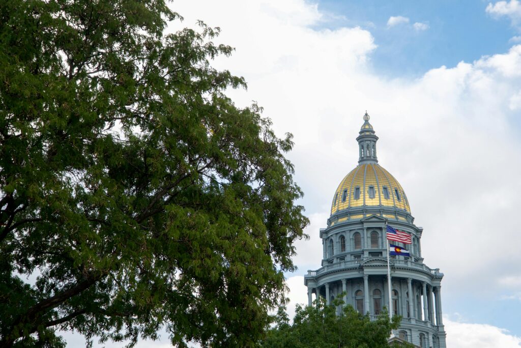 The Colorado State Capitol building with its iconic gold-domed roof, partially framed by a large green tree on the left. The U.S. flag and Colorado state flag are flying on flagpoles in front of the building, set against a partly cloudy blue sky.