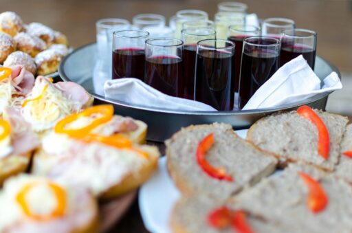 A buffet table featuring a variety of appetizers and drinks. In the foreground, open-faced sandwiches topped with sliced red peppers, cheese, and other ingredients are displayed on plates. In the background, a tray holds glasses of red wine and clear beverages, placed on a white cloth. Powdered pastries are partially visible to the side.