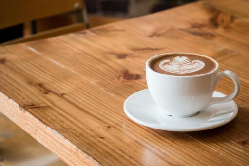 A white ceramic cup of hot coffee with latte art in the shape of a leafy pattern, placed on a matching white saucer on a wooden table with a natural finish.