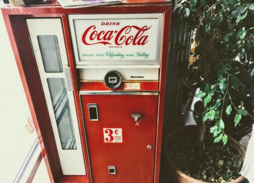 A vintage Coca-Cola vending machine with a red and white design, displaying the slogan 'Enjoy that Refreshing New Feeling.' The machine features a coin slot, bottle opener, and a price of 3 cents prominently displayed.